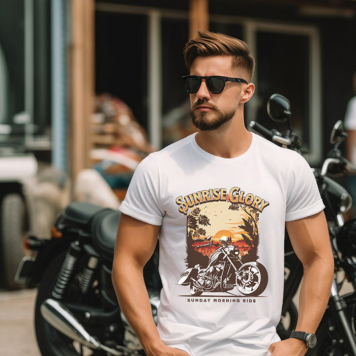 A man wearing a white streetwear t-shirt featuring a vintage-style graphic of a cruiser motorcycle parked in front of a tropical sunset with palm trees. The words 'SUNRISE GLORY' are printed in a bold, arched yellow font at the top, and 'SUNDAY MORNING RIDE' is written in clean black text at the bottom standing in front of motorcycles.